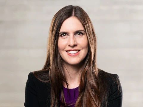 Frau mit langen Haaren in schwarzem Blazer vor neutralem Hintergrund.