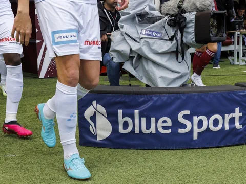 Fussballspieler laufen an Kamera mit 'blue Sport'-Logo vorbei.