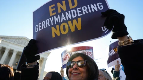 Person hält ein Schild mit der Aufschrift 'End Gerrymandering Now' vor einem Gebäude.