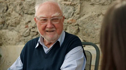 Älterer Mann mit Brille sitzt draussen und blickt auf Person im Vordergrund.