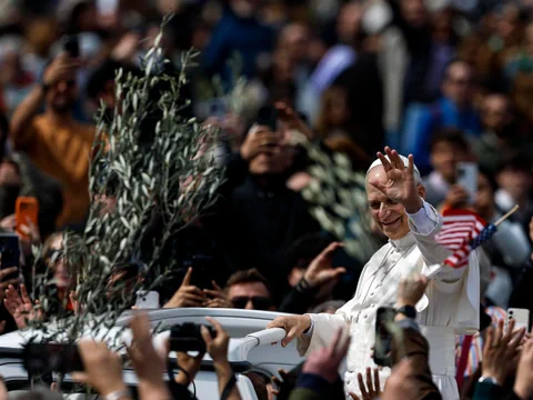 Papst Leo XIV. in weissem Gewand winkt, umgeben von Menschenmenge mit Fahnen und Zweigen.