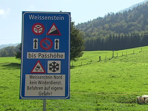 Strassenschild vor grüner Wiese und bewaldetem Hügel.