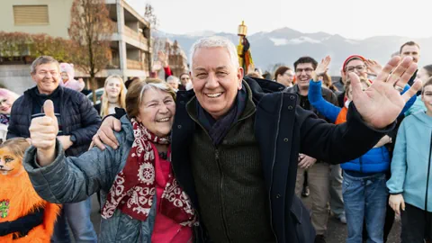 Links eine Frau und rechts ein Mann winken in die Kamera. Hinten ihnen stehen viele Leute.