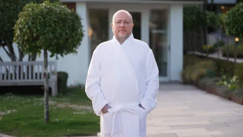 Mann in Bademantel steht im Garten vor einem Gebäude.