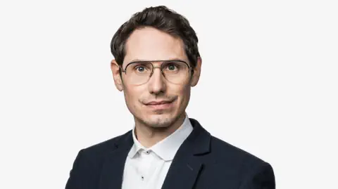Mann mit Brille im Anzug vor weissem Hintergrund.
