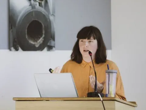 Person hält Vortrag am Rednerpult mit Laptop und Getränk.