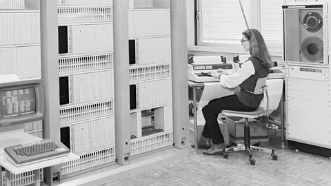 Person mit Brille sitzt an einem Schreibtisch vor alter Computertechnik.