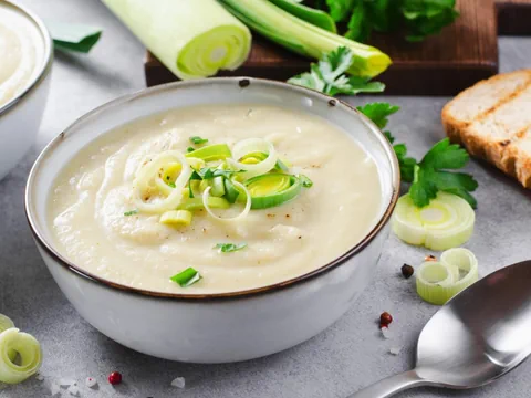 Schale mit Cremesuppe, garniert mit Lauch, Löffel und Brot daneben.