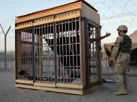 Mensch in Käfig mit Soldat daneben im Freien.