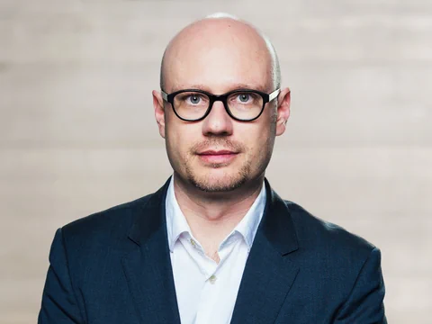 Mann mit Brille und Anzug vor neutralem Hintergrund.