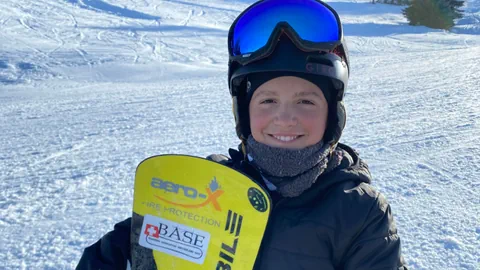 Gaël hält sein Snowboard in der Hand, steht auf der Piste und grinst in die Kamera