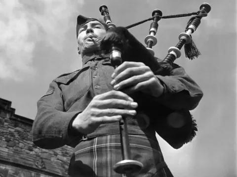 Mann in traditioneller Kleidung spielt Dudelsack vor Steinmauer.