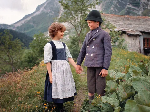 Ein Mädchen und ein Junge in traditioneller Kleidung stehen in einer Berglandschaft und halten sich an den Händen.