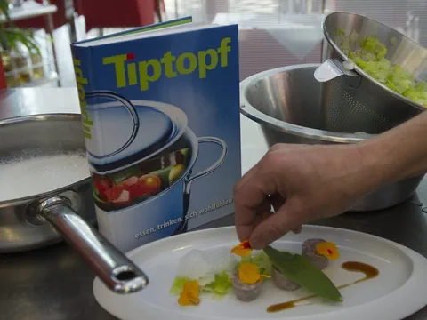 Person dekoriert Teller mit Tiptopf-Kochbuch im Hintergrund.