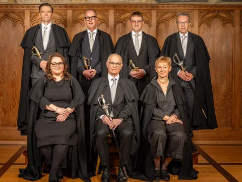 Gruppe von Personen in Roben mit Schwertern in einem Holzgetäfelten Raum.