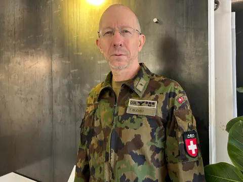 Mann in Tarnuniform mit Abzeichen, steht vor schwarzem Hintergrund.