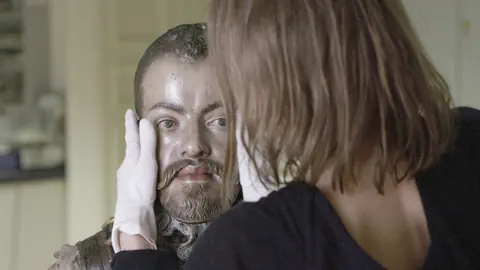 Eine Frau hält mit weissen Handschuhen den Kopf einer Skulptur, die beinahe wi ein echter Mann aussieht.  