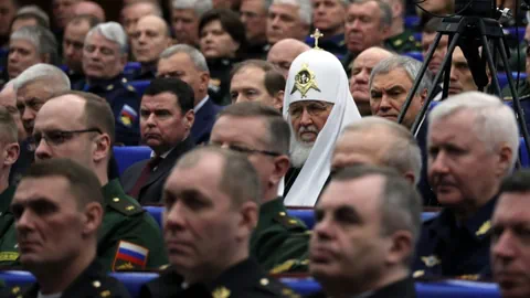 Patriarch Kirill ist in kirchlichem Gewand zwischen uniformierten Militärs zu sehen.
