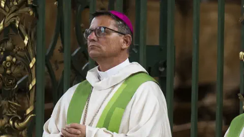 Priester mit Brille und lila Käppchen vor grünem Hintergrund.