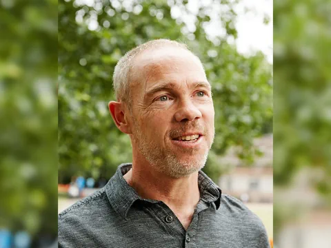 Ein Mann mit grauem Hemd im Freien mit Bäumen im Hintergrund.