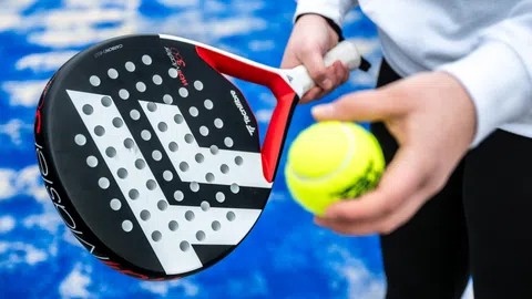 Person hält einen Padel-Schläger und Ball auf blauem Platz.