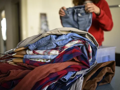 Person hält gefaltete Jeans, Kleidung liegt auf Tisch.
