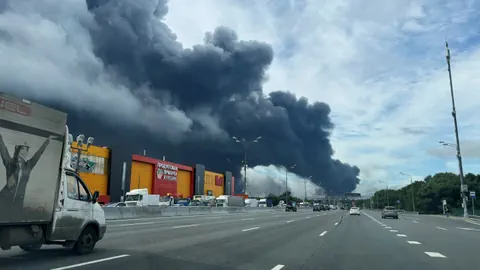 Strasse mit vorbeifahrenden Fahrzeugen und grossen Rauchwolken am Himmel.