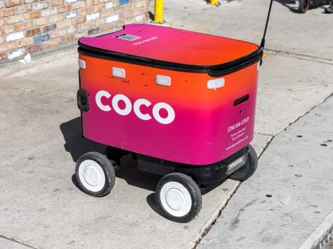 Kleiner Lieferroboter mit rosa-orangefarbener Oberfläche auf Gehweg.