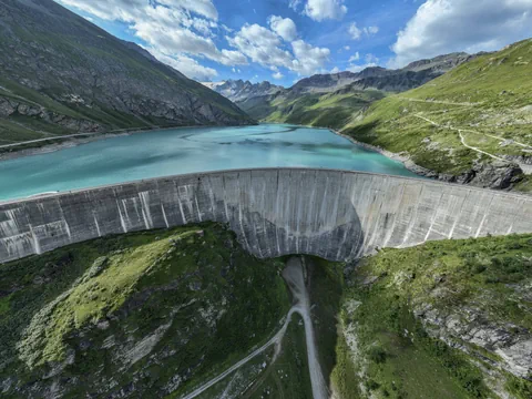 Grosser Staudamm mit türkisfarbenem See und umliegenden Bergen unter blauem Himmel.
