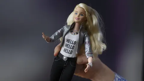Blonde Puppe in silberner Jacke und 'Hello' Shirt, in Hand gehalten.