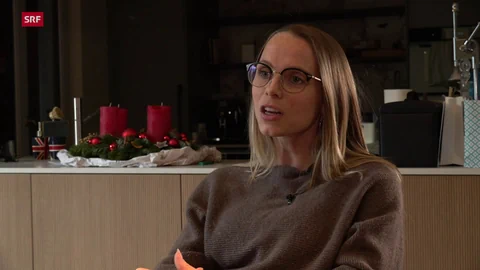 Frau mit Brille spricht in einem Raum, Kerzen und Weihnachtsdekoration im Hintergrund.