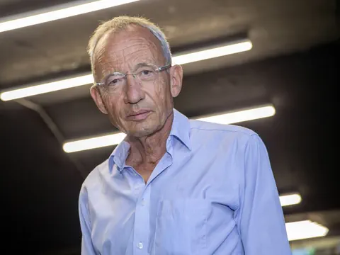 Älterer Mann mit Brille und blauem Hemd steht vor dunklem Hintergrund mit mehreren horizontalen Lichtern.