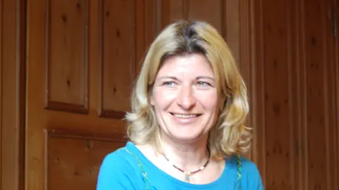 Zu sehen ist das Gesicht einer Frau mittleren Alters. Sie hat blond gefärbte Haare, trägt eine Halskette und ein türkis-farbenes Oberteil. Sie lächelt. Dem Anschein nach ist sie amüsiert. Oder glücklich.