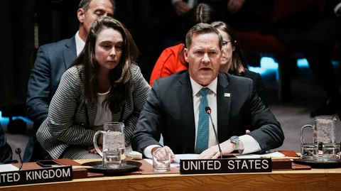 Personen sitzen bei einer öffentlichen Veranstaltung an einem Tisch mit 'United States'-Schild.