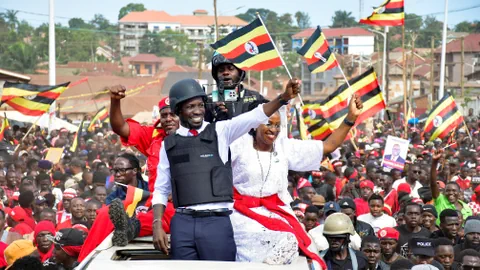 Bobi Wine steht durch das offene Dach eines Autos und winkt mit einer ugandischen Flagge der Menschenmenge um das Fahrzeug zu. Er trägt eine Schutzweste und einen Helm.