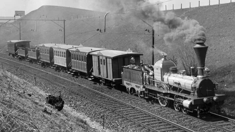 Eine Schwarzweiss-Aufnahme eines Dampfzuges. Die Lokomotive stösst Rauch aus. 