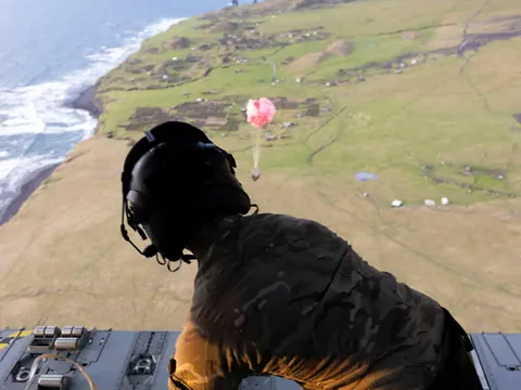 Soldat schaut von Flugzeug über Küstenlandschaft, abgeworfene Fracht mit Fallschirm.
