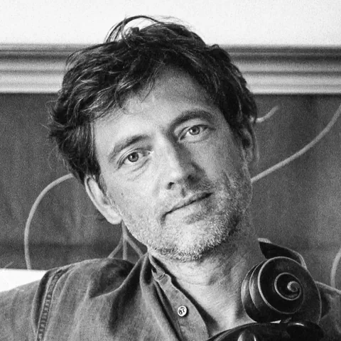 Schwarz-Weiss-Fot eines Mannes mittleren Alters mit Dreitagebart und weichem Blick. Vor ihm ein Cello.