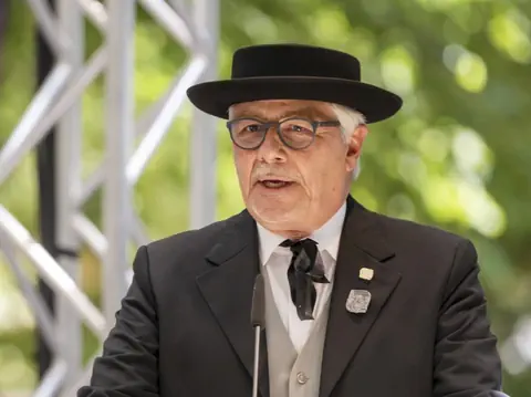 Älterer Mann mit Brille und Hut spricht im Freien, grüne Bäume im Hintergrund.