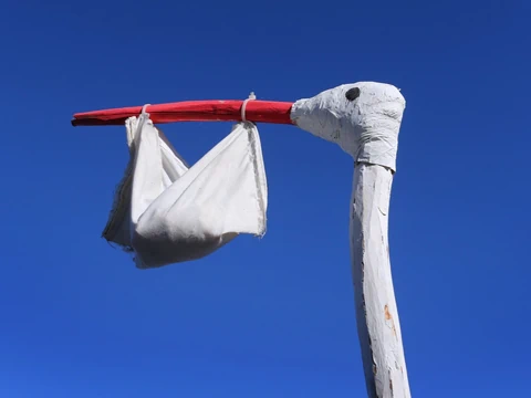 Storchenskulptur mit Stoffbündel im Schnabel vor blauem Himmel.