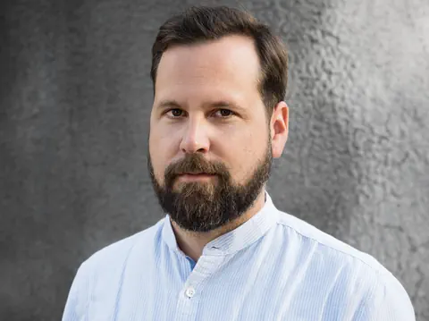 Mann mit Bart und Hemd vor grauem Hintergrund.