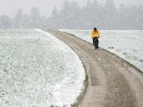 Mann fährt Fahrrad auf einer verschneiten Strasse neben Feld.