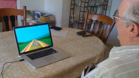 Älterer Herr sitzt vor einem Laptop, auf diesem sieht man ein Spiel mit einem Auto.