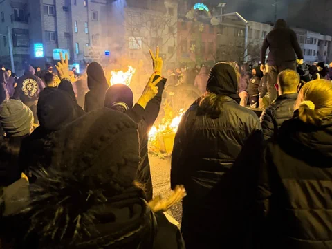Menschenmenge bei Nachtprotest mit brennendem Feuer im Hintergrund.