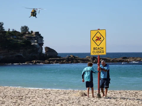Zwei Personen und ein geschlossenes Strand-Schild mit Helikopter im Hintergrund.