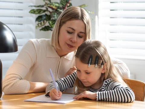 Frau hilft Kind beim Schreiben am Tisch.