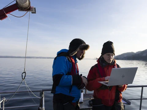 Zwei Personen auf einem Boot arbeiten am Laptop auf einem See.