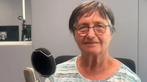 Ältere Person mit Brille lächelt vor einem Mikrofon in einem Studio.
