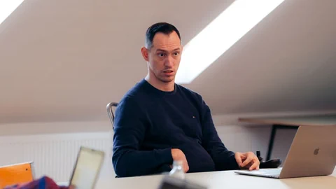 Islam Alijaj sitzt vor einer Dachschräge an einem Tisch mit einem geöffneten Laptop.