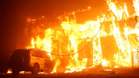 Ein Gebäude in Flammen, davor steht ein Auto. 